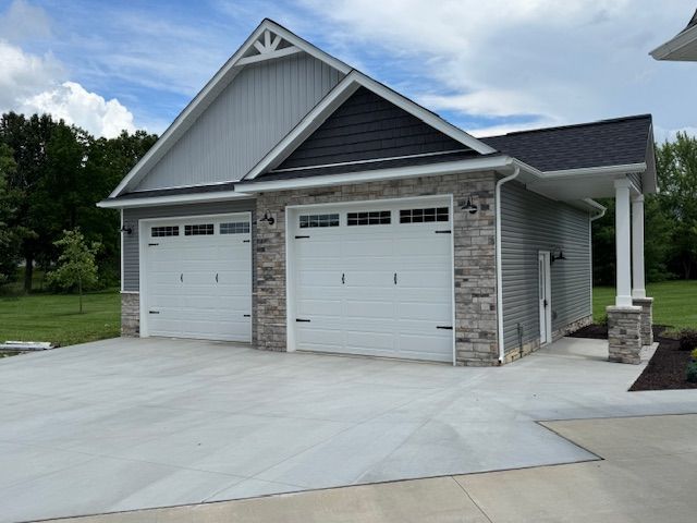 Two-car garage with gray siding and white doors, front driveway, and small covered porch under cloudy sky
