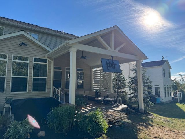Covered porch of a suburban house with hanging sign, bright sun, and landscaped front yard.