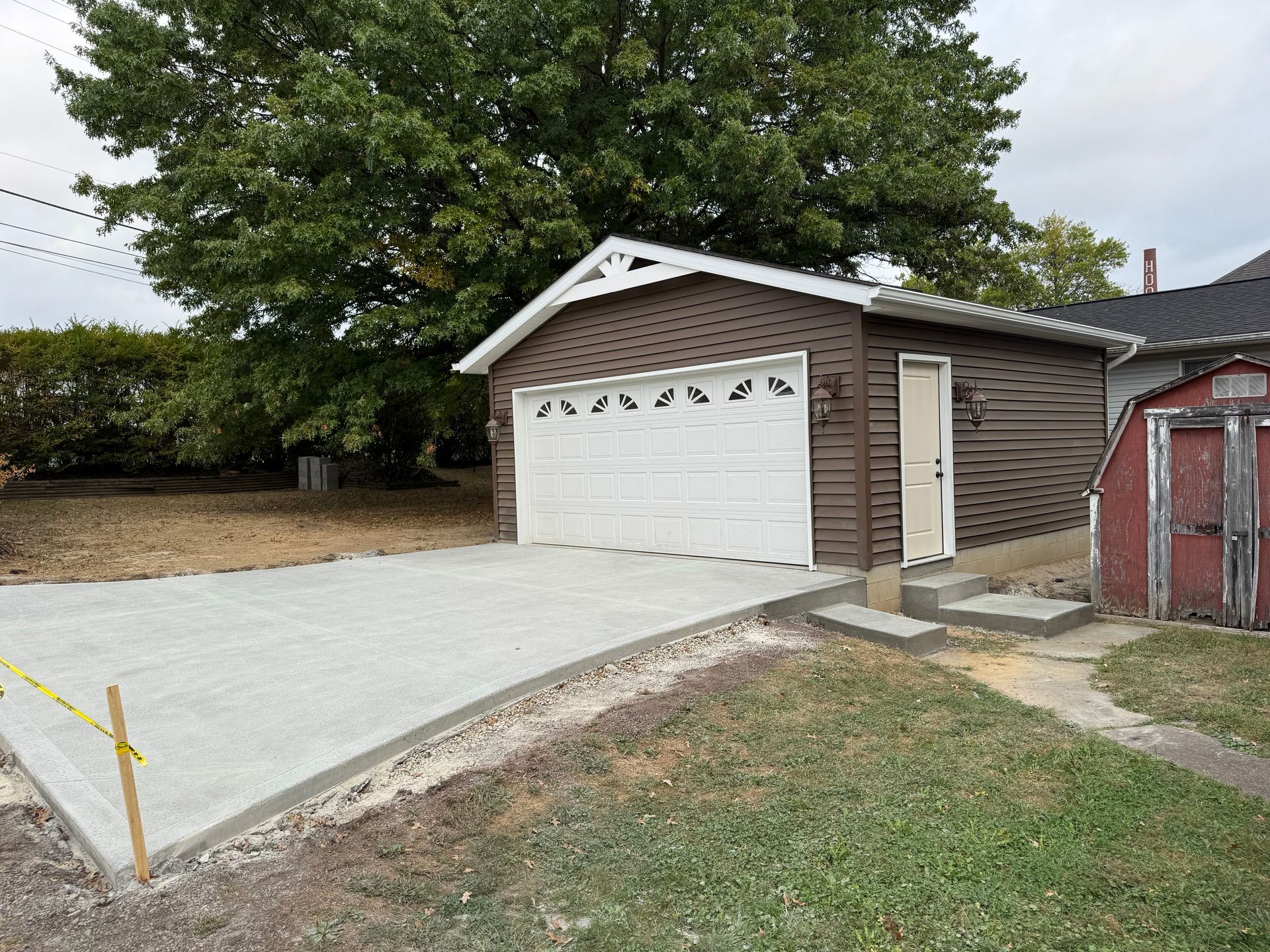 Detached two-car garage with white doors, concrete driveway, and a large tree in front of a house yard