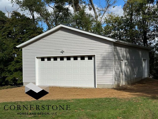 White detached garage with a single white door and beige siding, surrounded by grass and trees.