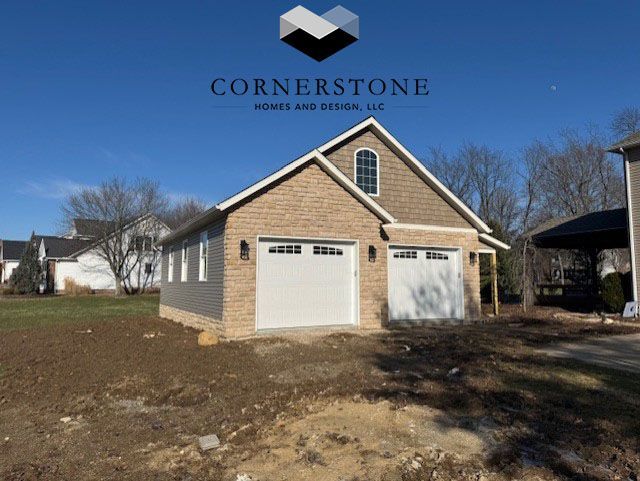 Brick garage with two white doors and tan siding under a blue sky, branded Cornerstone Homes & Construction logo above