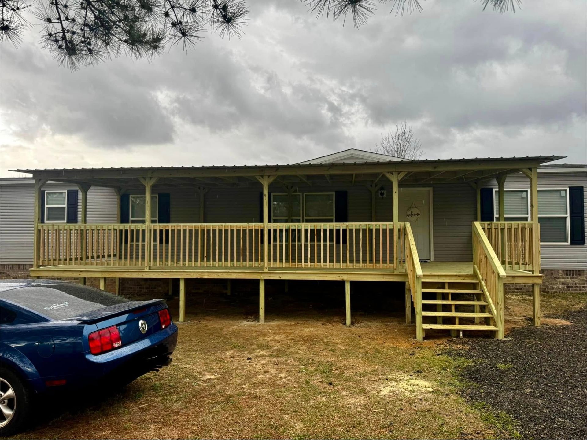 Blue car parked next to a light-colored mobile home with a large wooden porch. Cloudy sky.