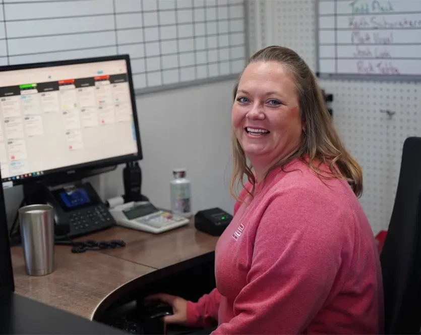 Woman smiling at desk with computer, phone, and white schedule board.