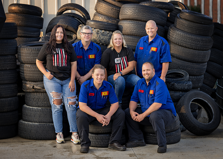 Group of six people in blue work shirts pose in front of a tire pile.