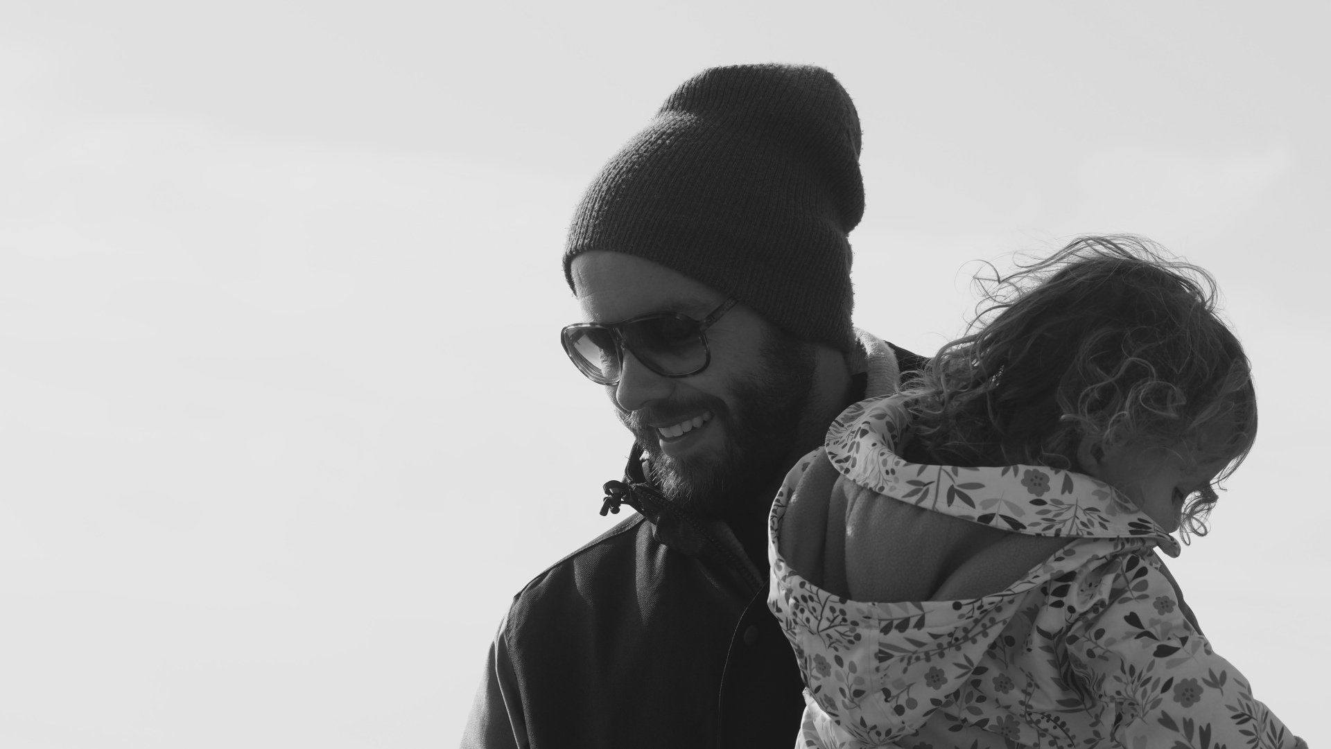 Smiling person in a beanie holding a bundled child against a bright sky