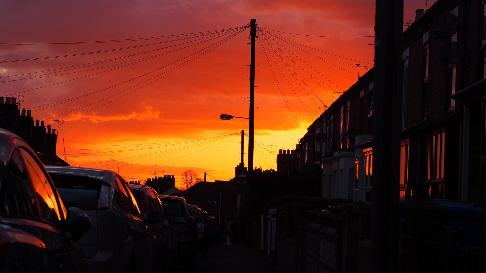 Sunset over a residential street with silhouetted houses, parked cars, and a utility pole against a fiery orange sky