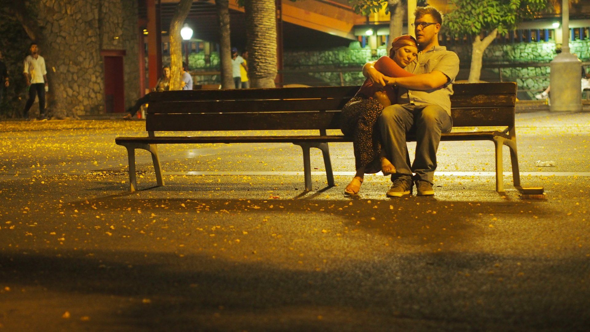Person sitting on a park bench at night under warm streetlights, surrounded by fallen leaves.