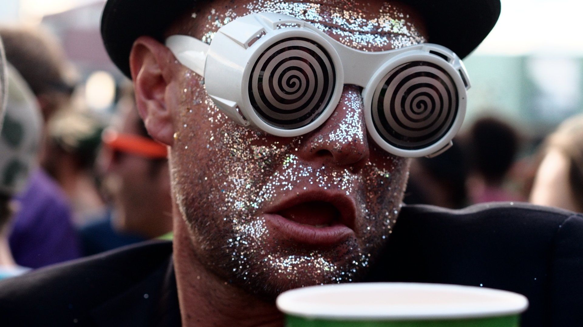 Person in spiral glasses and sparkly face paint at a crowded event, holding a cup