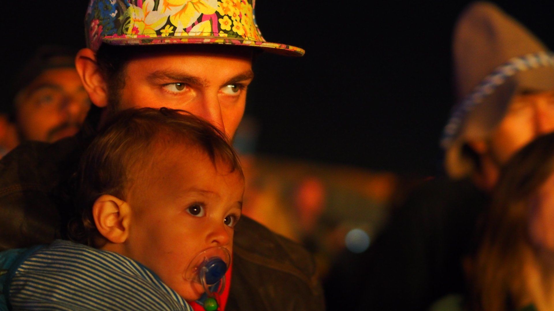 A child with a pacifier being held at a nighttime outdoor event, with a man in a colorful hat behind them