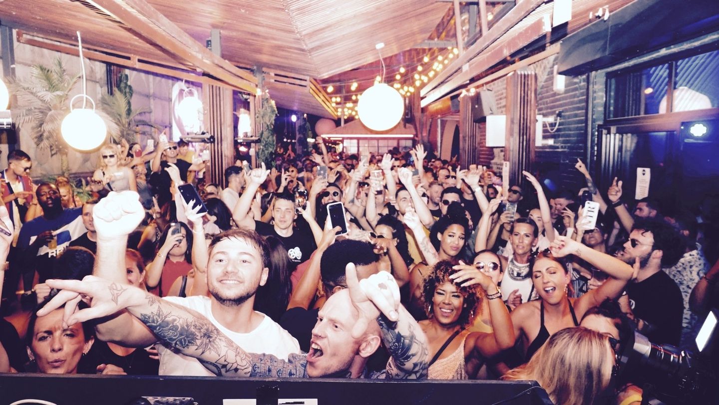 Crowded bar scene with people dancing and raising their hands under warm hanging lights
