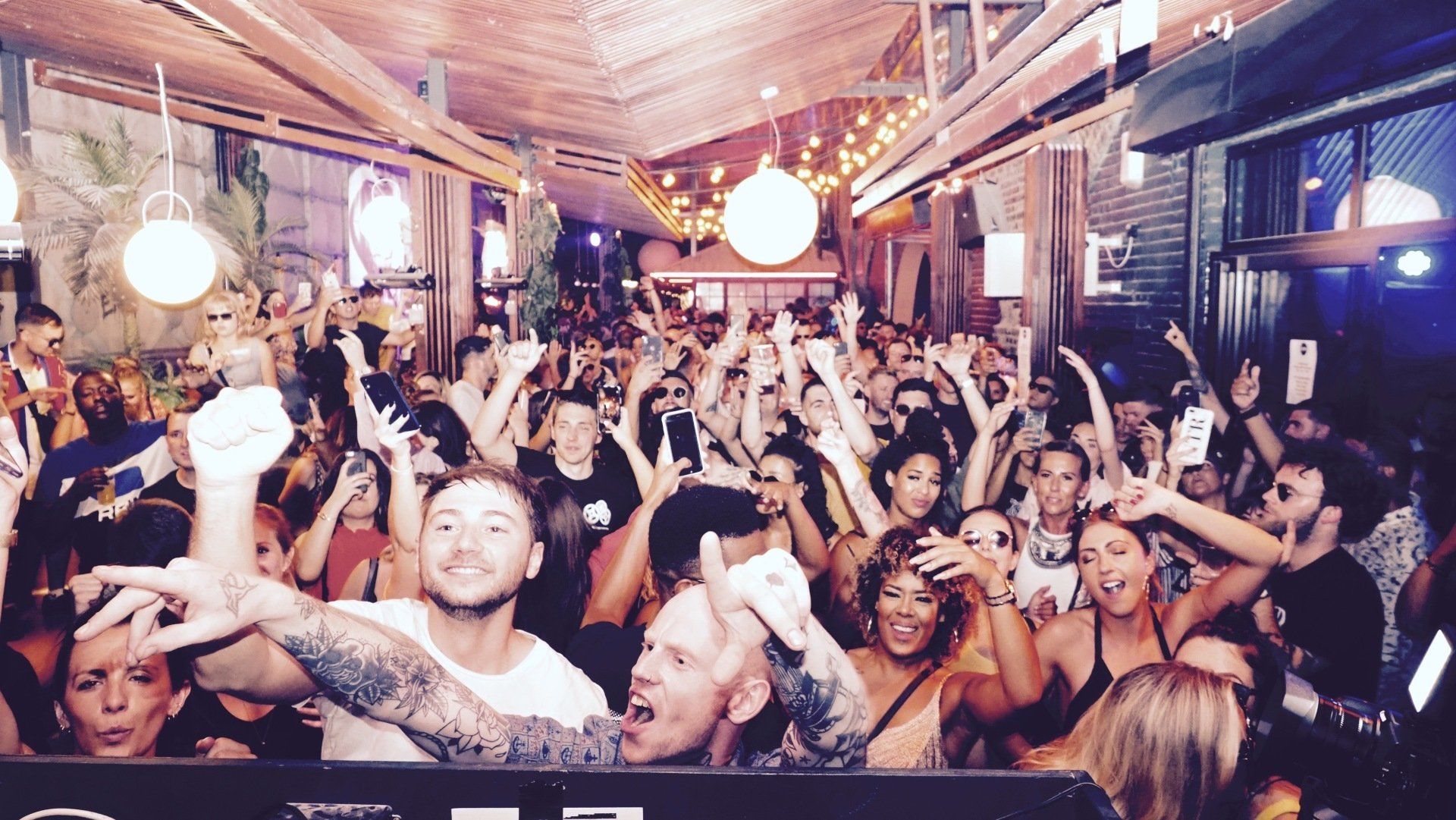 Crowded indoor party with people dancing, raising hands, and colorful lights in a warmly lit venue