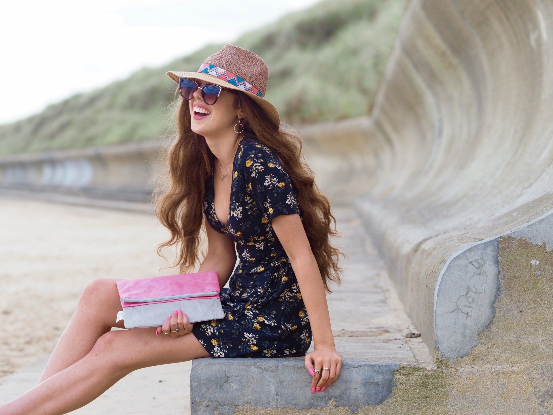 Woman in a floral dress and hat sitting by a curved concrete wall on a sandy path, smiling and holding a pink box
