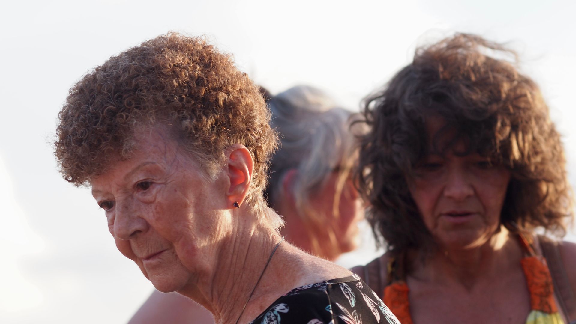 Two women with curly hair outdoors, one in foreground looking down, another behind in soft light.