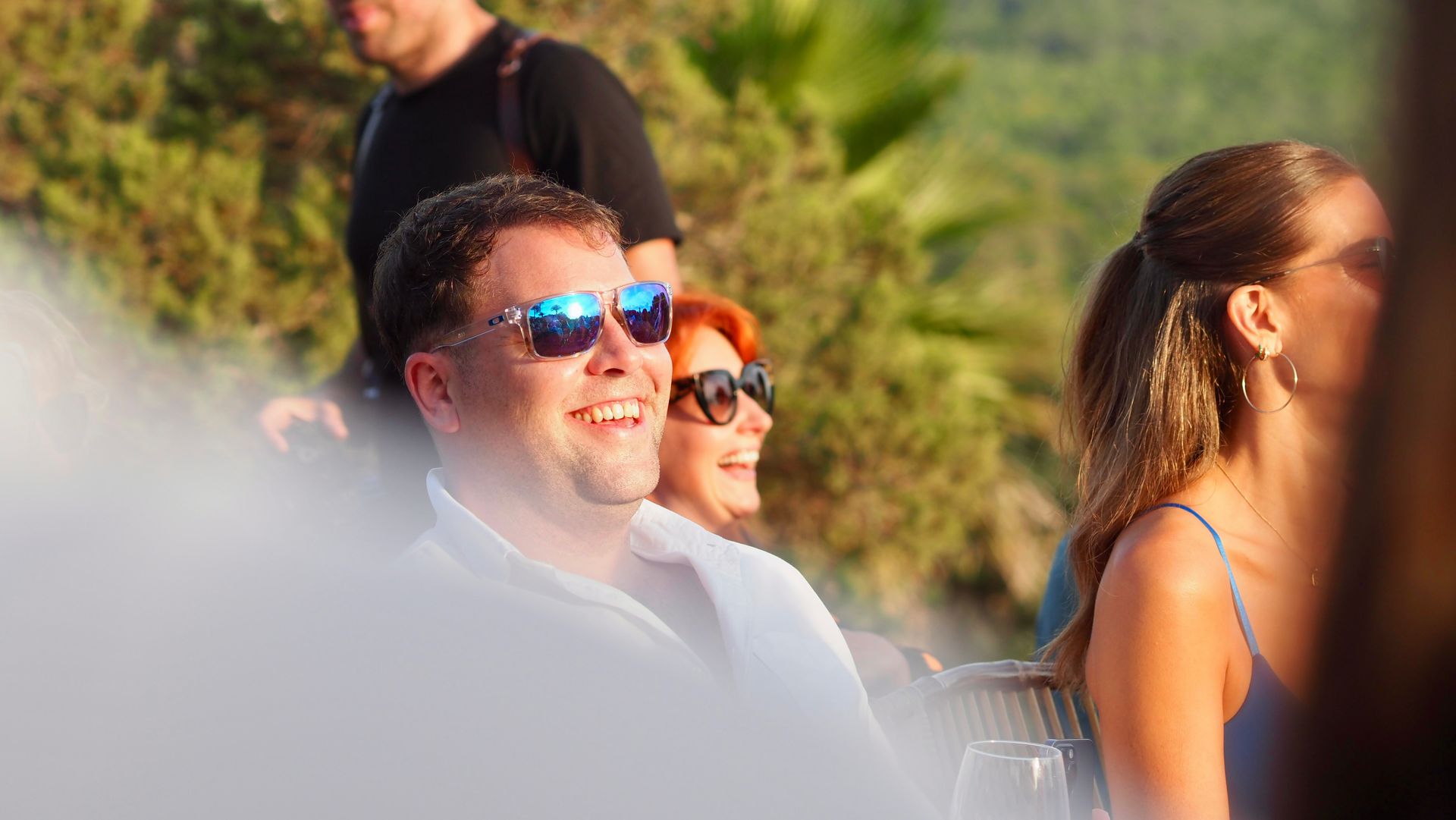 People seated outdoors in bright sunlight, including a man in blue reflective sunglasses and a woman in profile