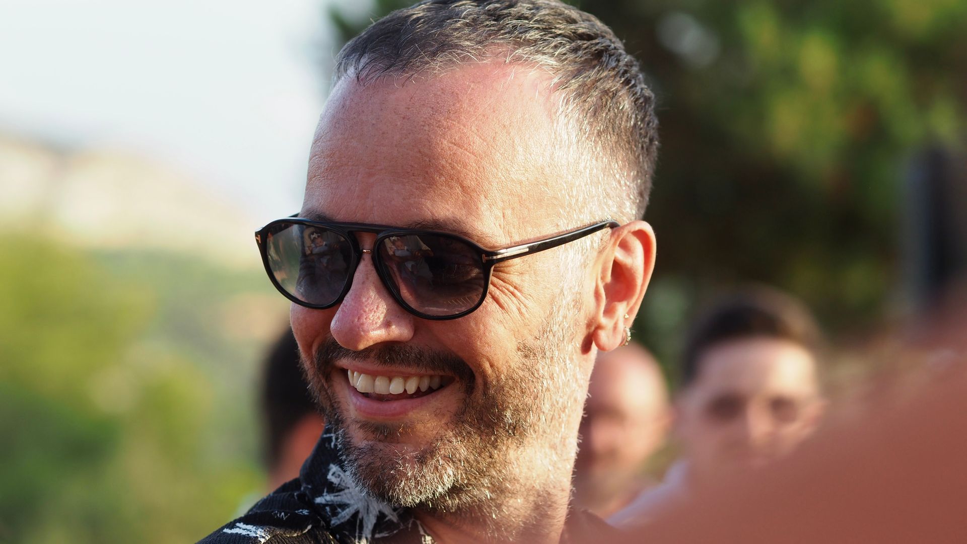 Smiling person wearing sunglasses outdoors with blurred people in the background