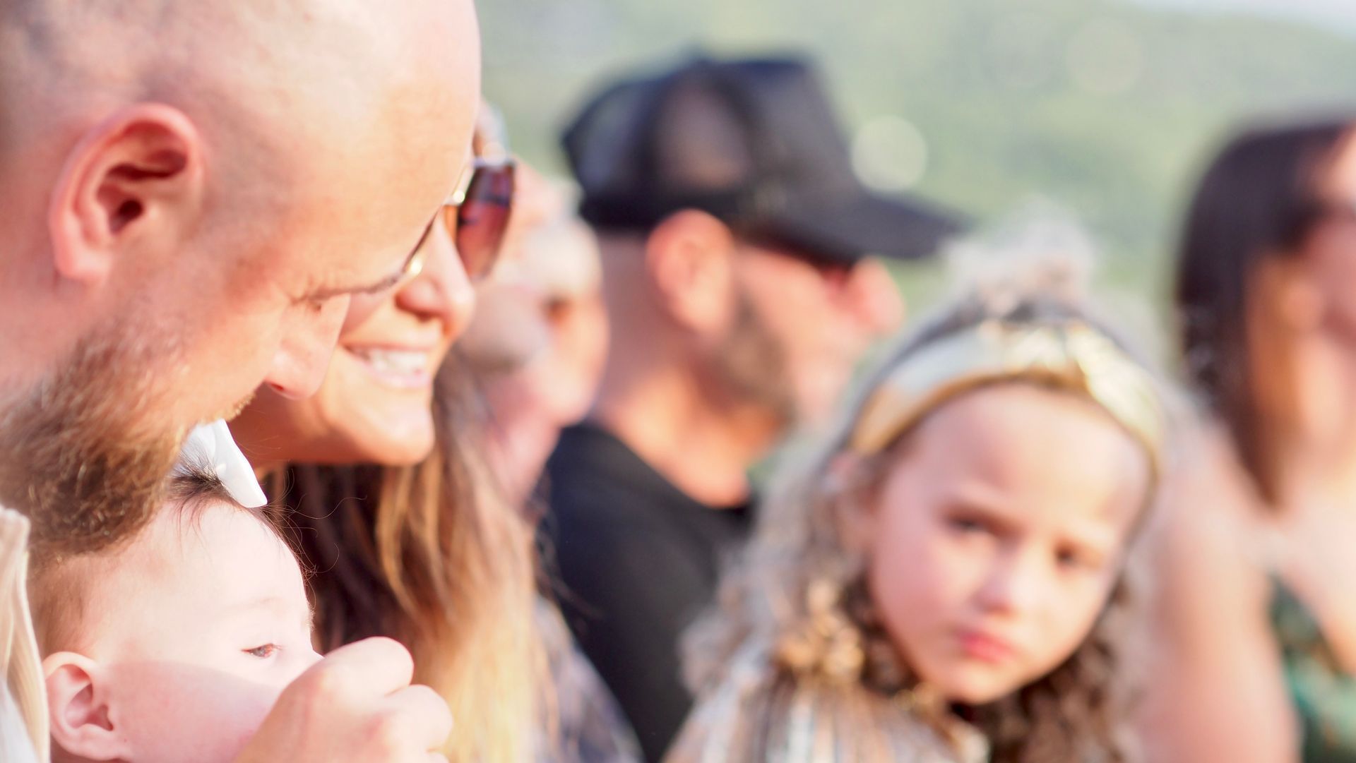 People standing outdoors in a crowd, with a bald man in the foreground and a child wearing a headband nearby.