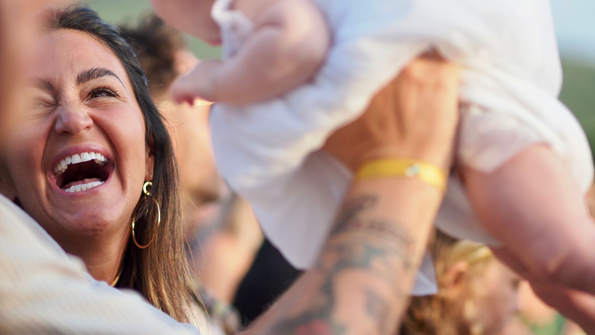 Smiling person at a celebration, holding up a baby in a white outfit, with a blurred crowd in the background