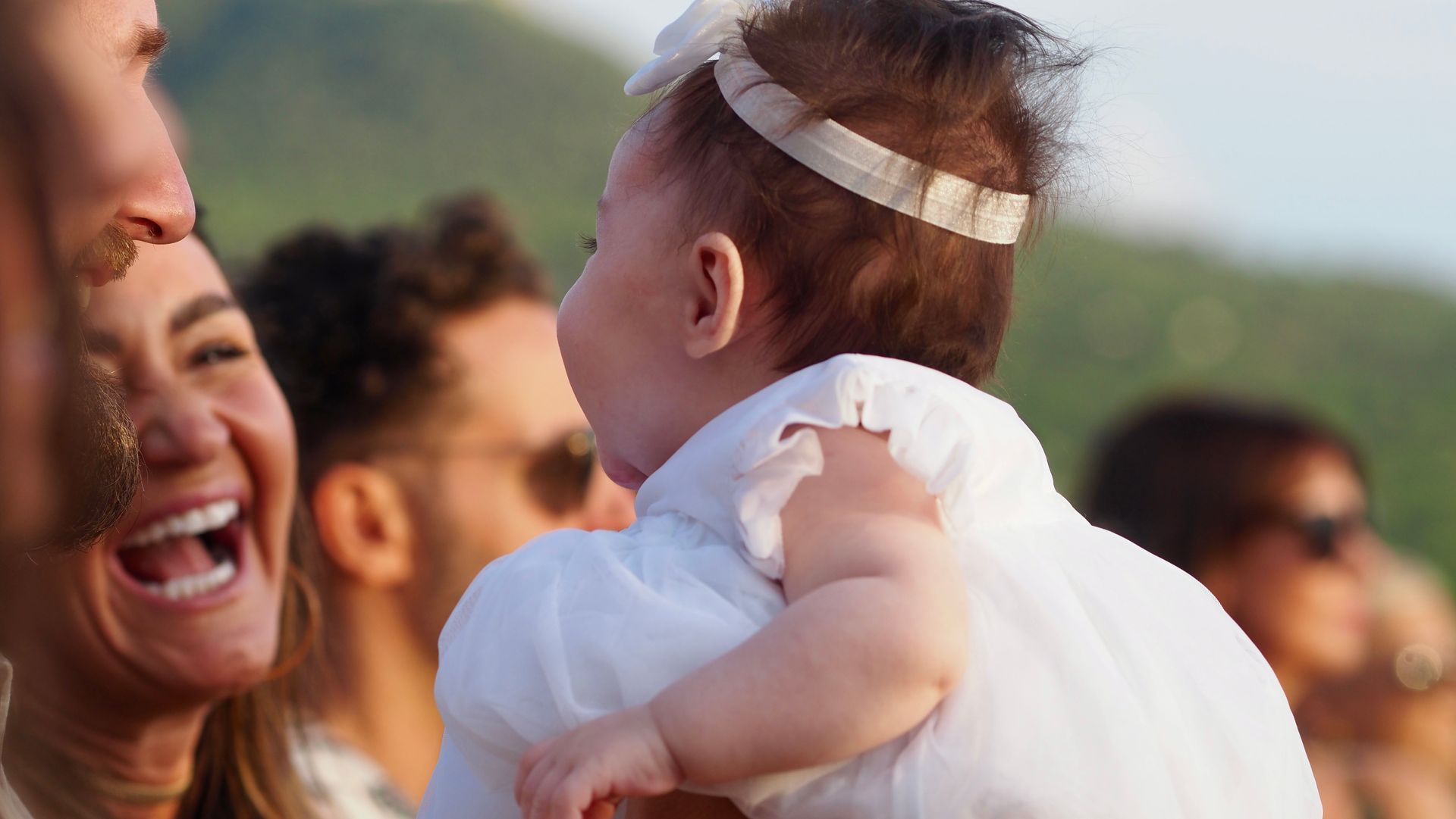 Smiling woman holding a baby at an outdoor gathering, with blurred people in the background.