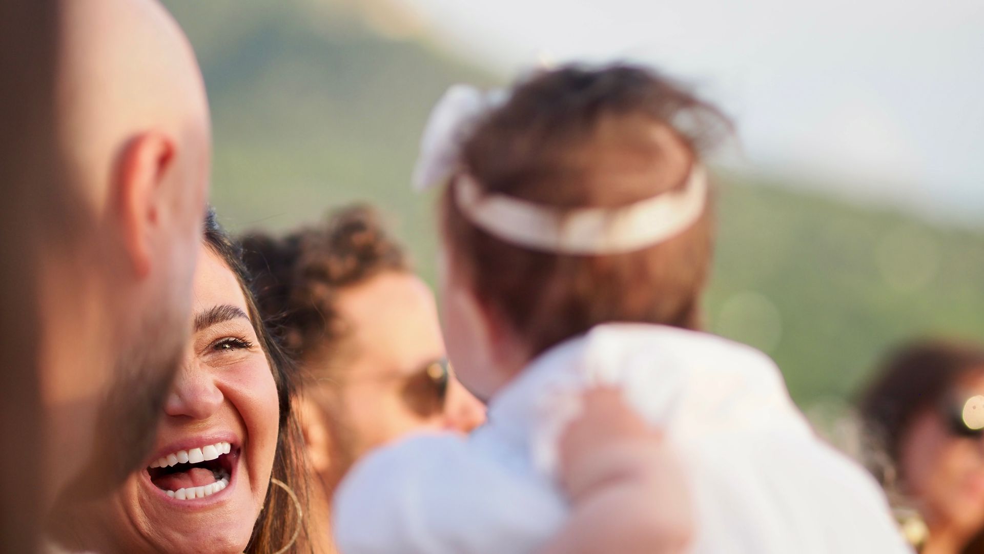 Smiling woman at an outdoor celebration, holding a child, with people blurred in the background