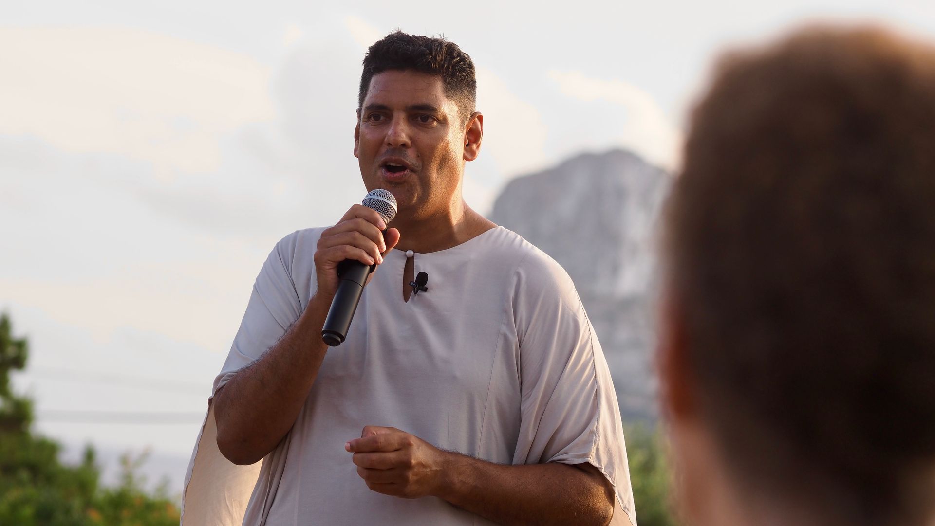 Man speaking into a microphone outdoors at sunset, with a blurred audience in the foreground.