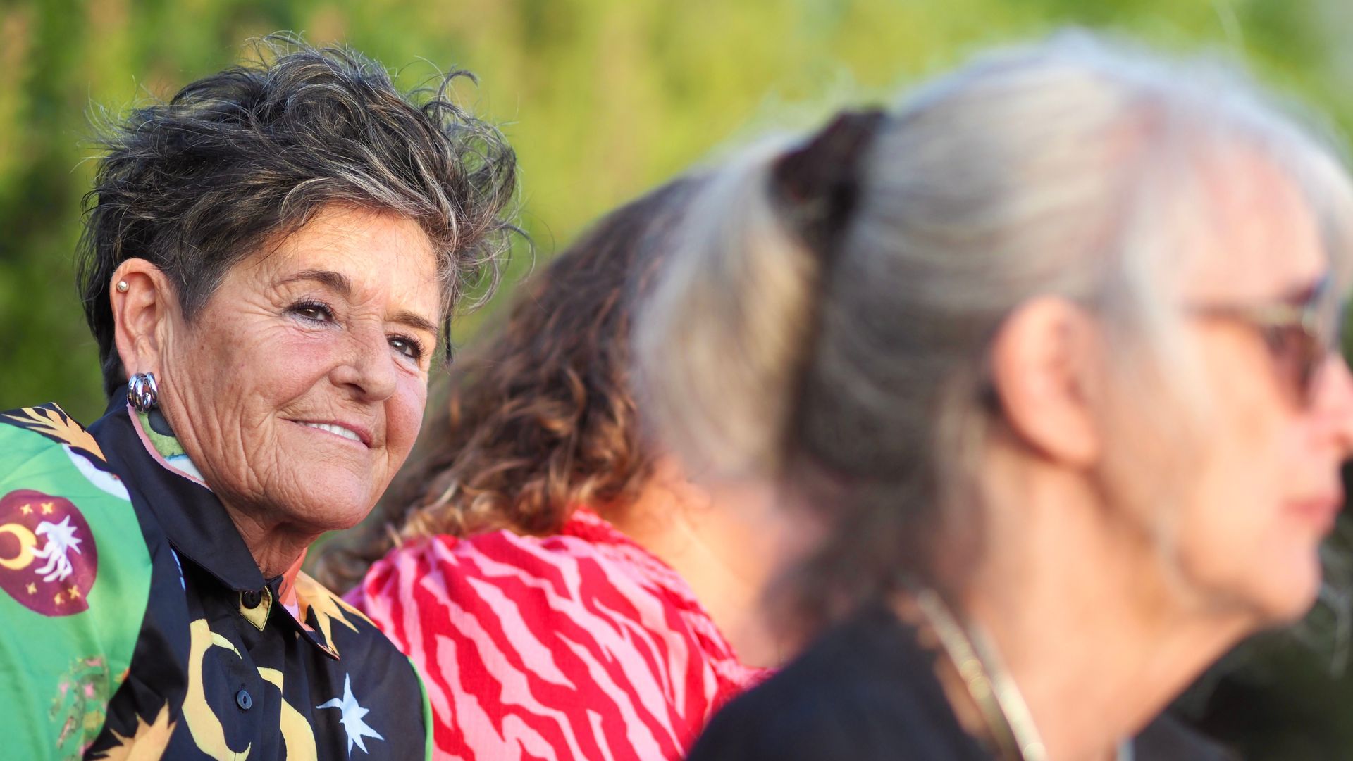Smiling woman in a floral shirt among seated people outdoors in bright sunlight