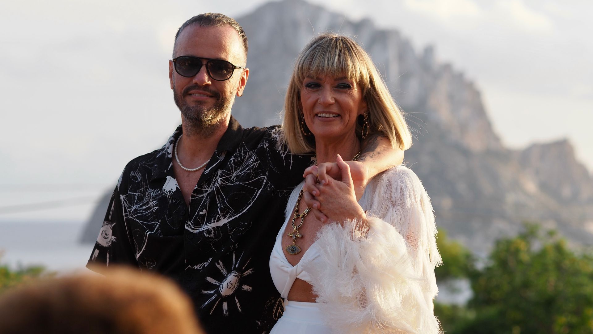 Two people posing outdoors in front of mountains, smiling in bright daylight.
