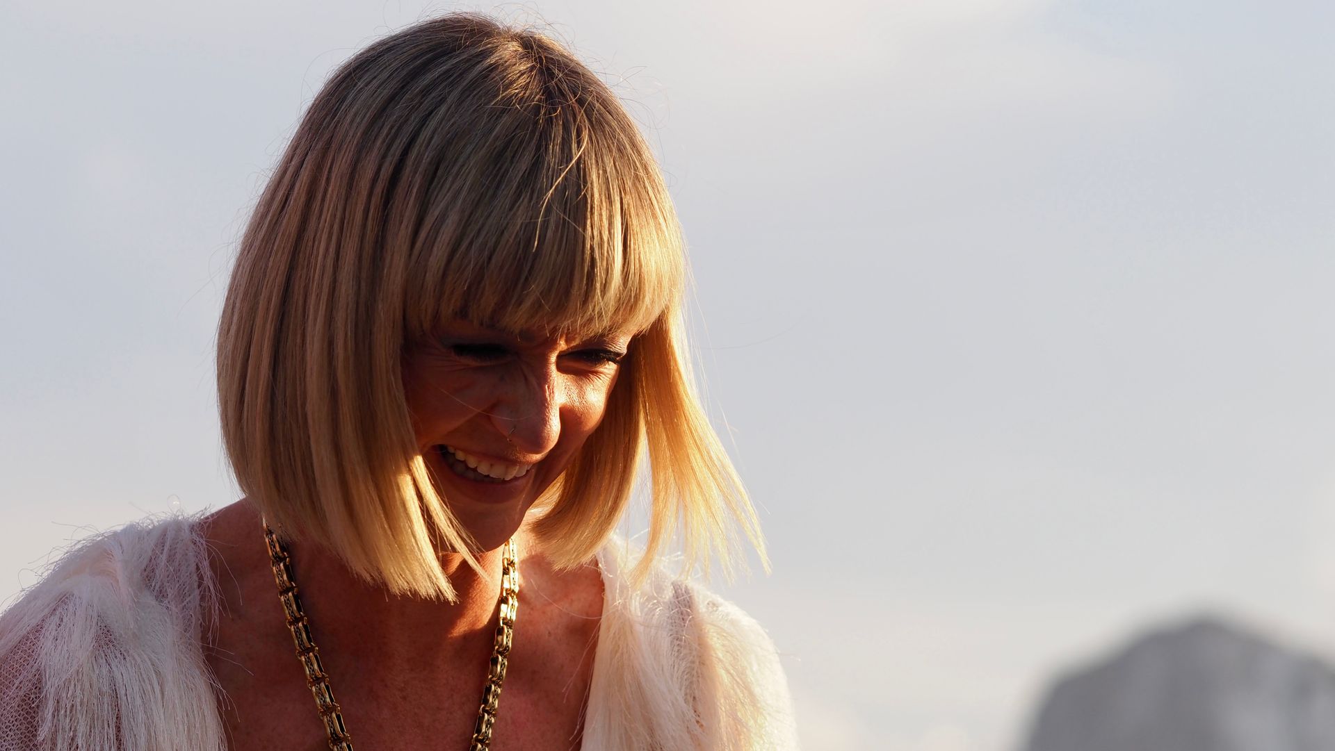 Smiling woman with short blonde hair looking down outdoors in warm sunlight