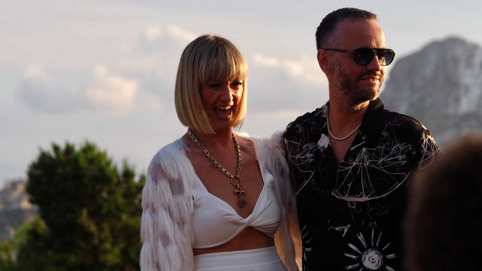 Two people standing outdoors at sunset, smiling and wearing white and black outfits.