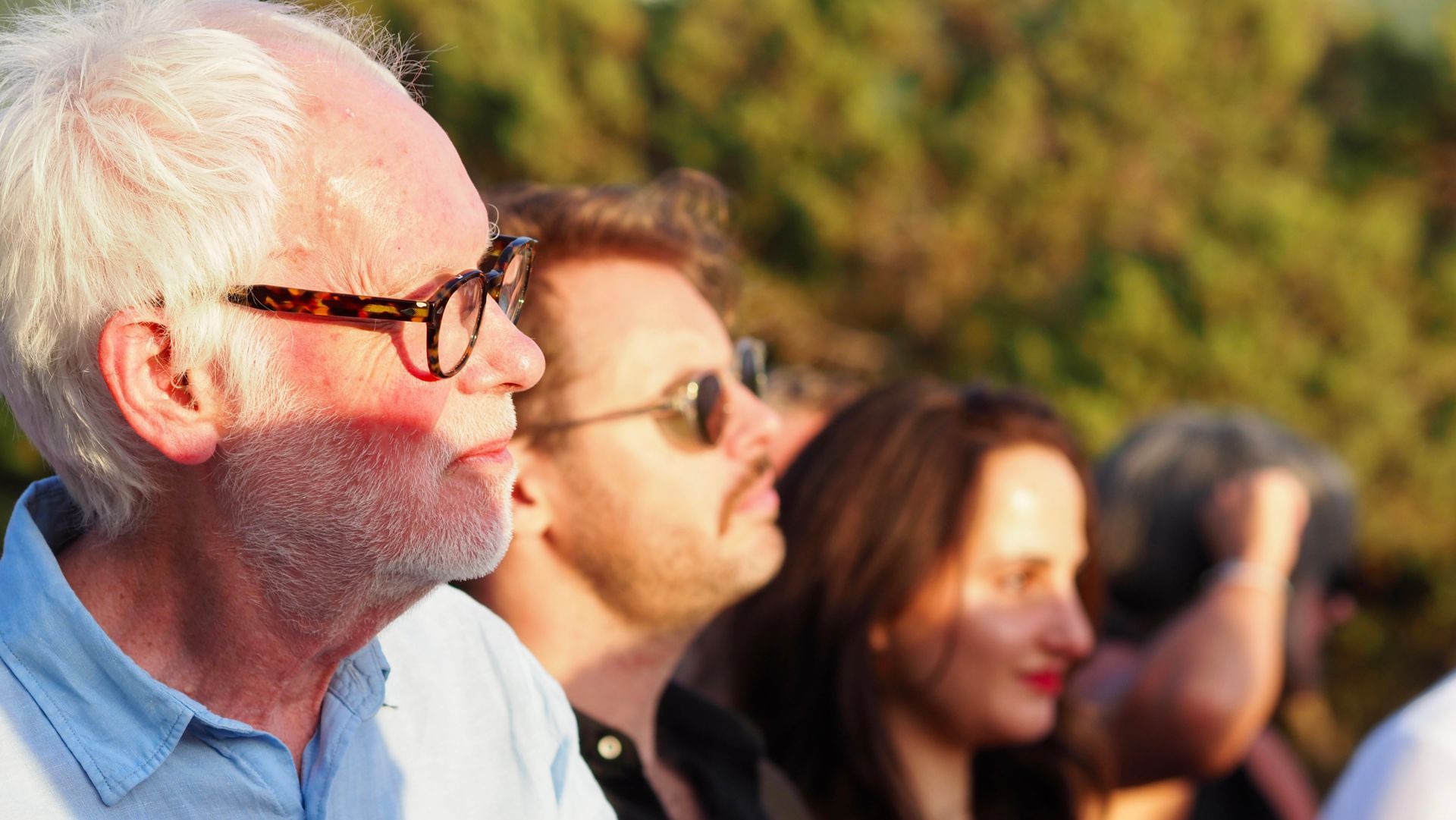 Close-up of an older man with glasses watching outdoors, with two blurred people in the background.