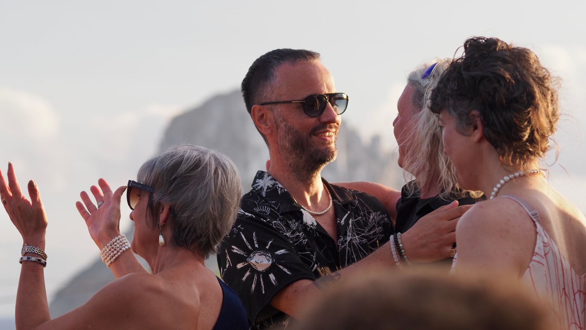 People dancing and socializing outdoors at a sunny event, with a man in sunglasses smiling in the center