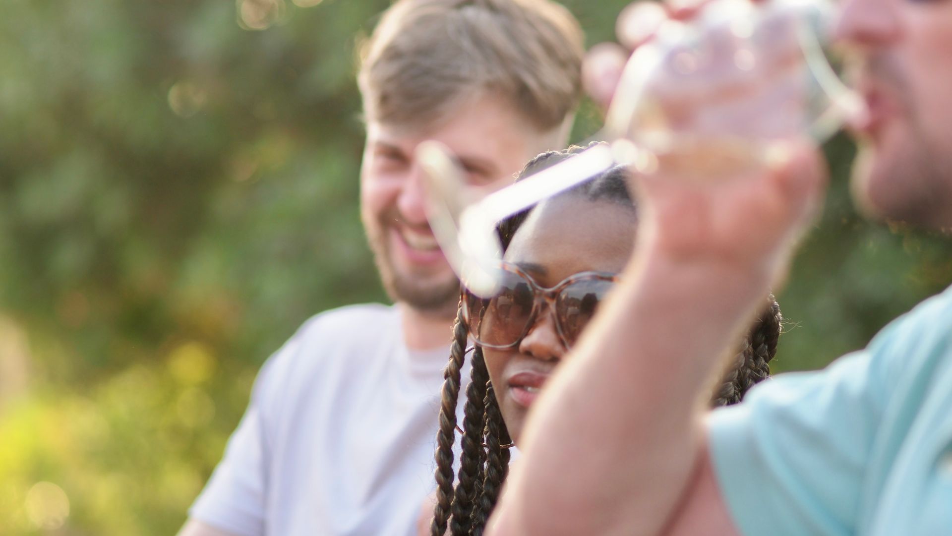 Three people outdoors, one holding up a drink, with a smiling person in sunglasses in the center
