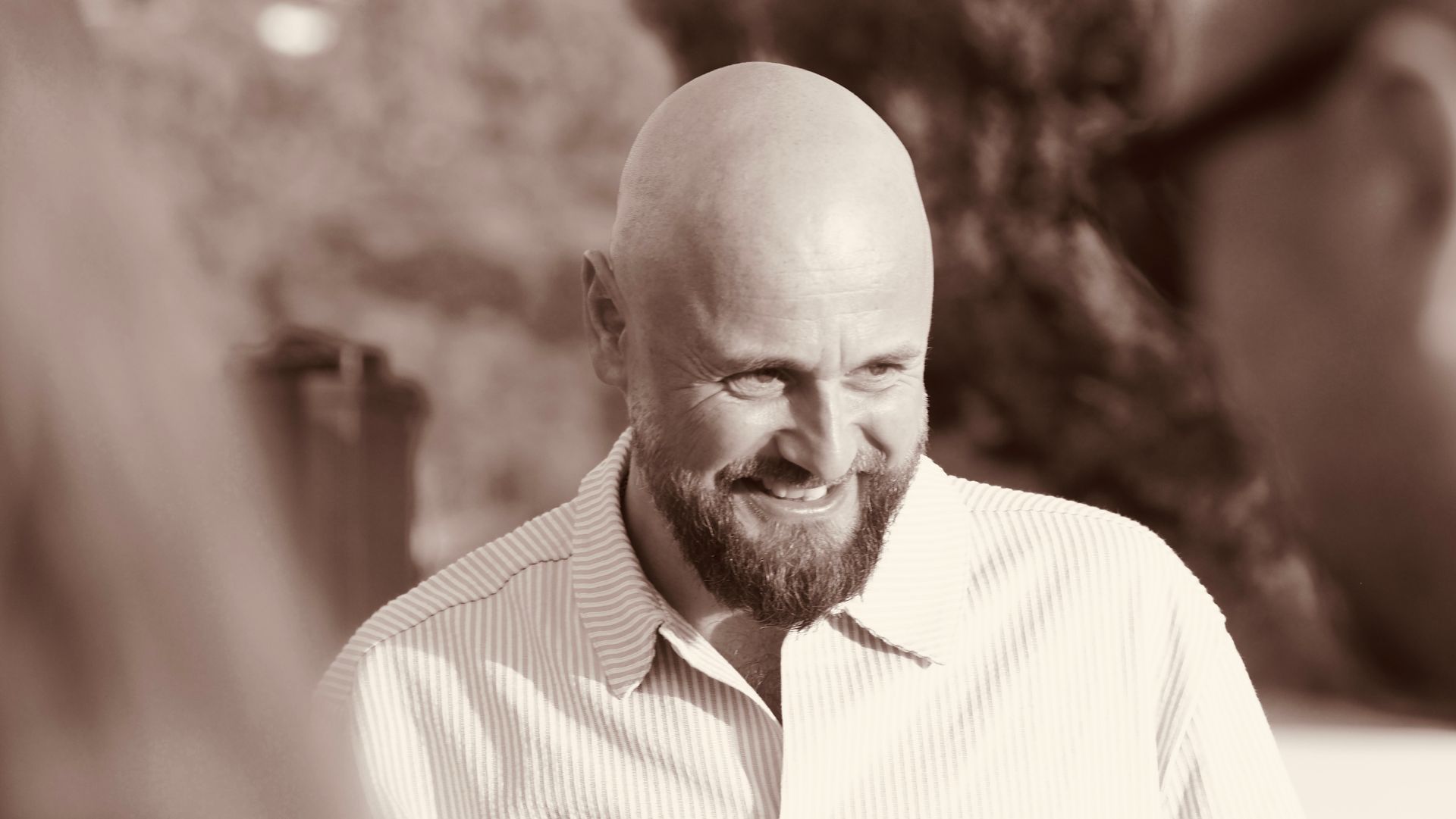 Smiling bald man in a striped shirt outdoors in warm, sepia-toned light