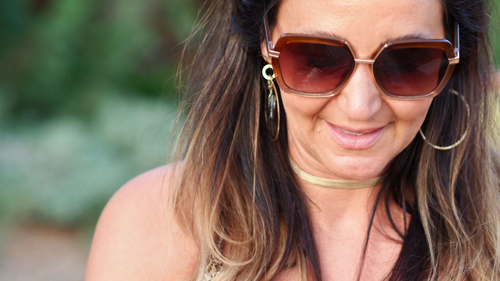 Woman wearing sunglasses and hoop earrings outdoors, smiling softly against a blurred green background