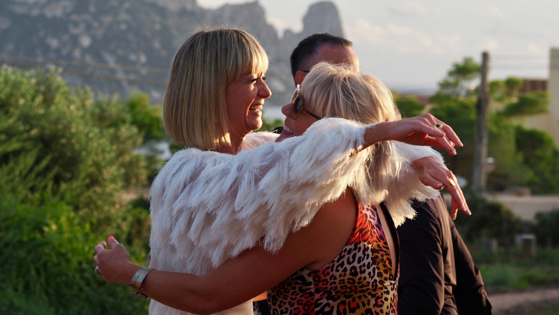 Two people embrace outdoors at sunset, with a third person nearby and mountains in the background