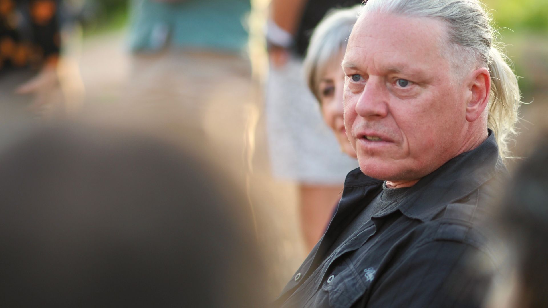 Close-up of a man with gray hair in a dark jacket, looking sideways with a serious expression outdoors.