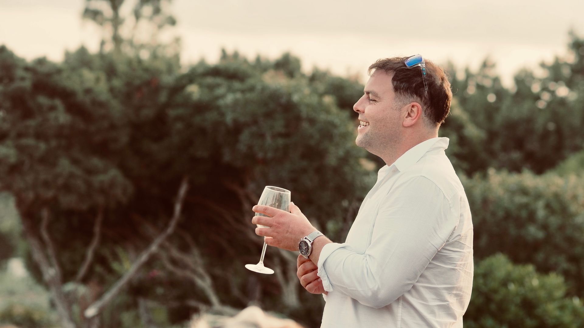 Man in white shirt holding a wine glass outdoors, smiling in front of greenery at sunset
