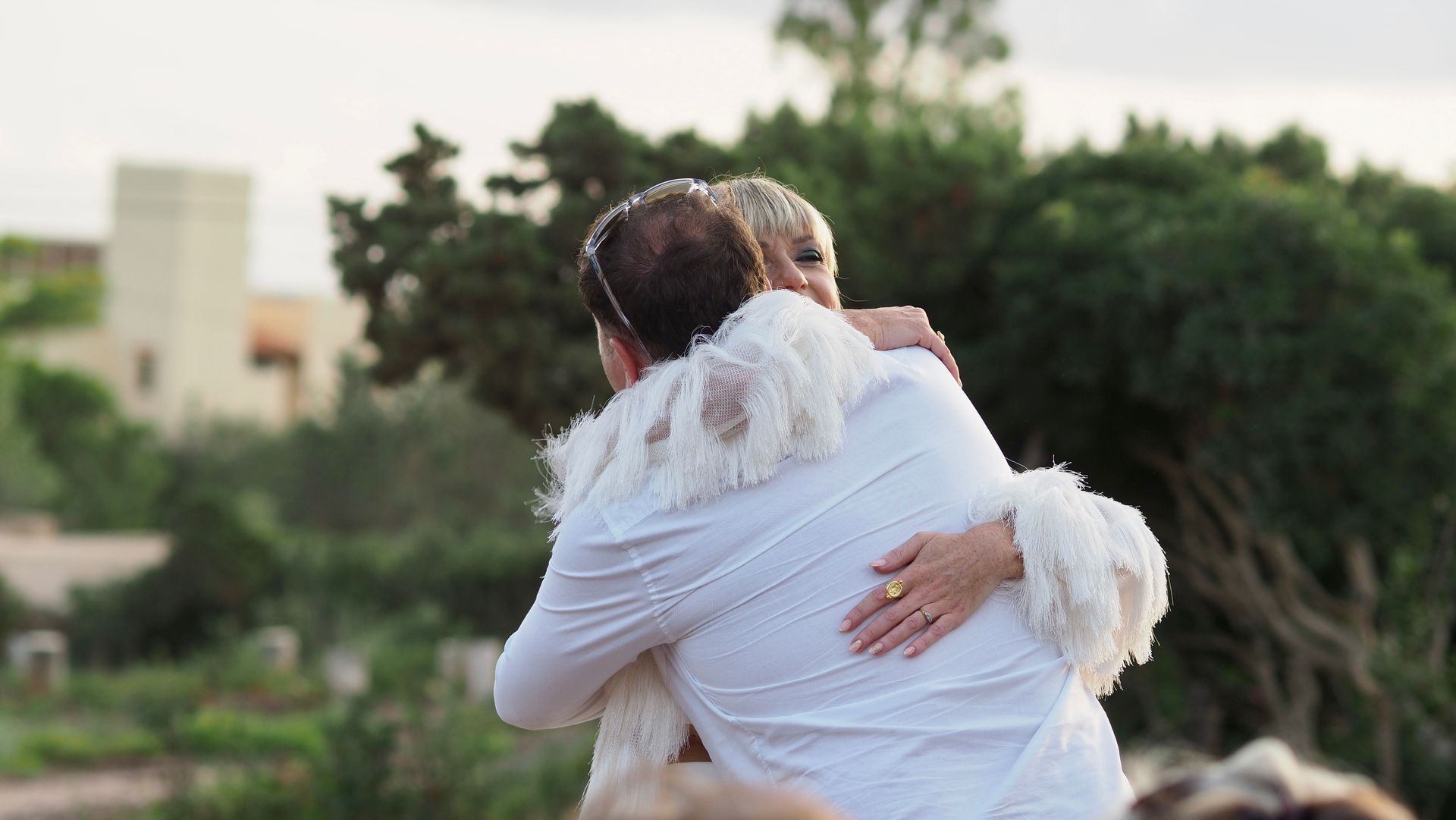 Two people in white clothing embrace outdoors with greenery in the background