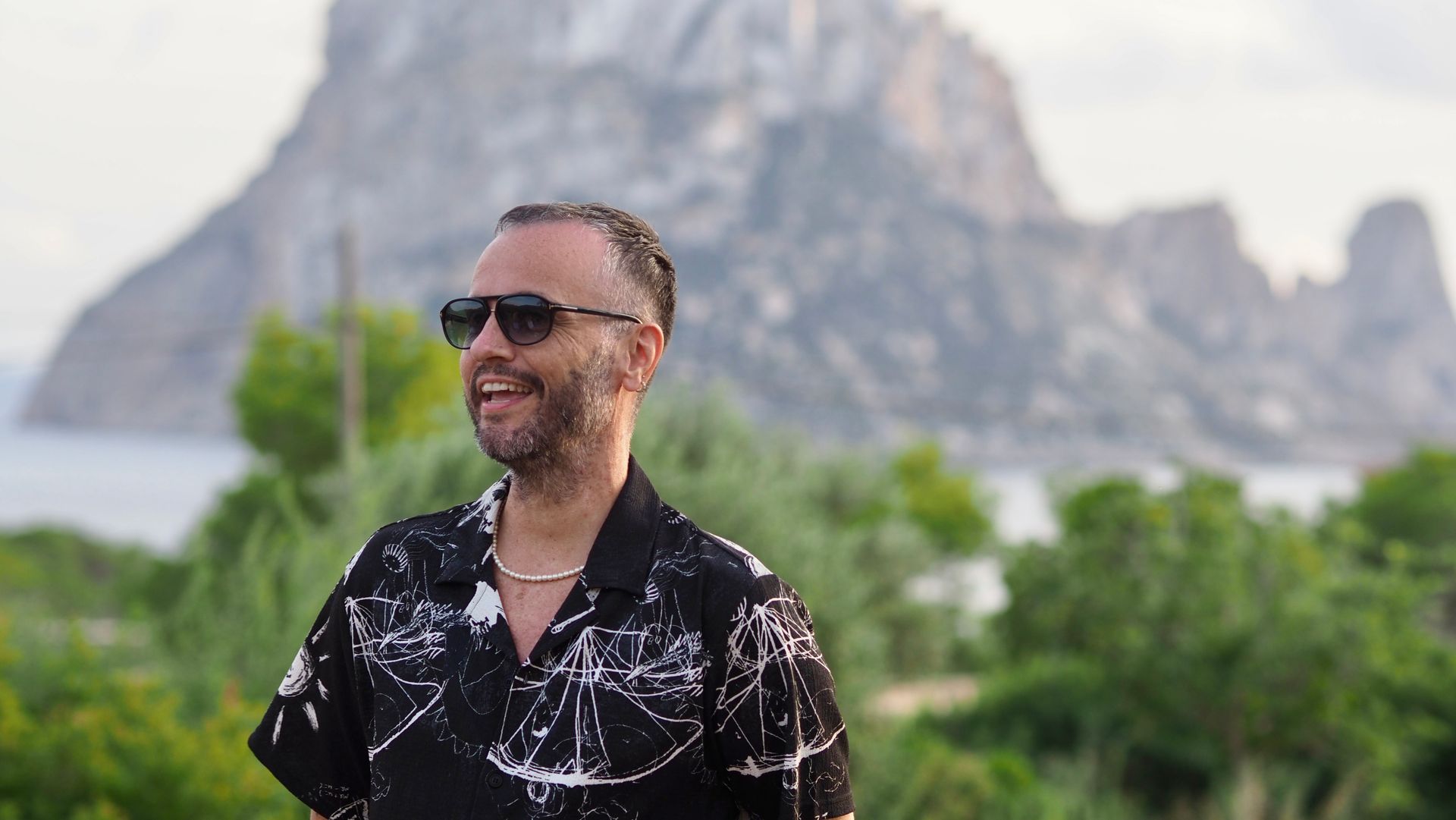 Man smiling in sunglasses and a black patterned shirt, standing outdoors with rocky mountains behind him.