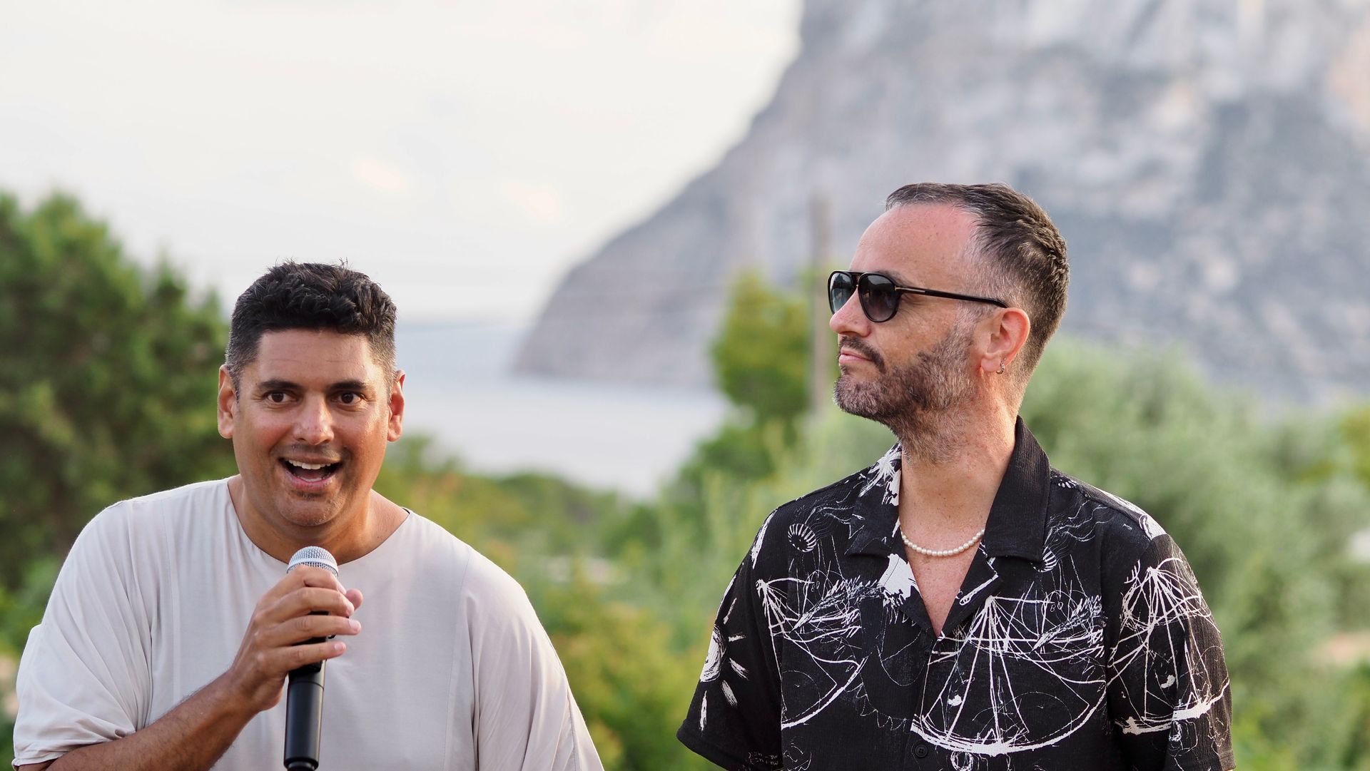 Two men outdoors, one holding a microphone, with mountains and greenery in the background.