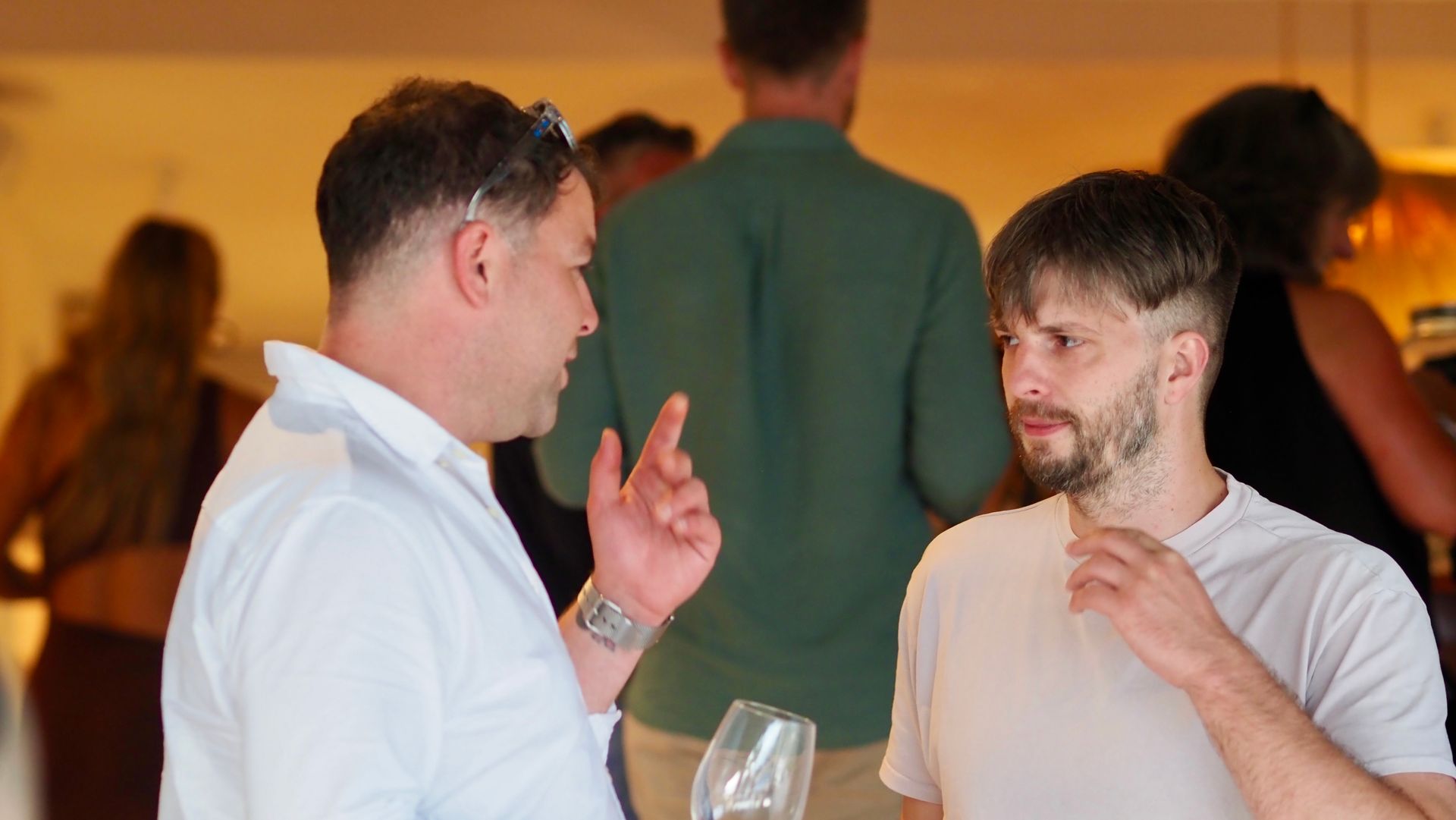 Two men talking at an indoor event, one gesturing while holding a drink, with blurred guests in the background.
