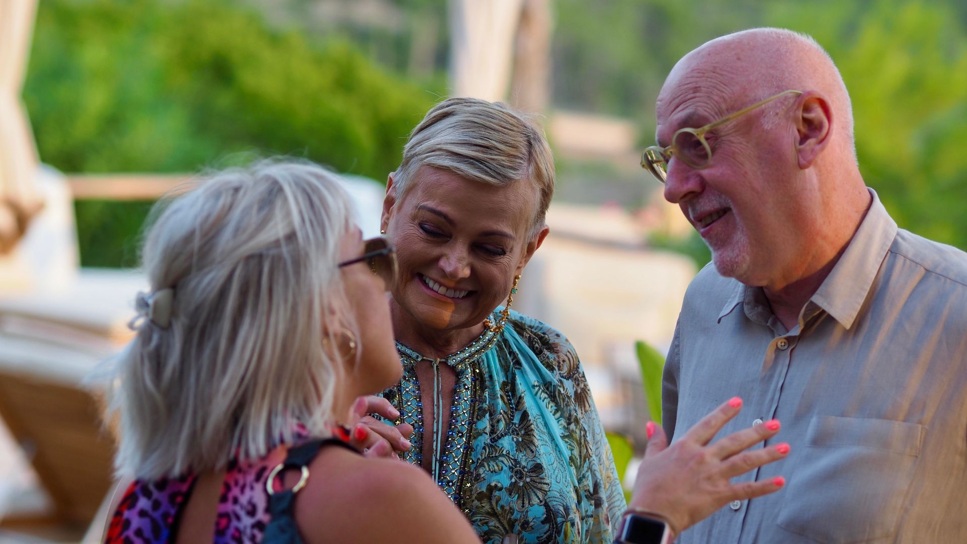 Three people smiling and chatting outdoors, with one woman gesturing as they stand in sunlight.