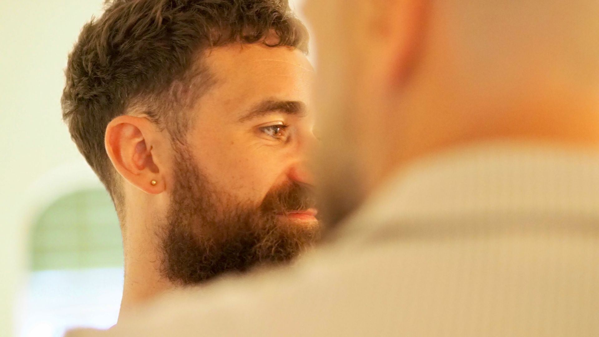 Two people in profile, one bearded man speaking closely to another in a bright indoor setting