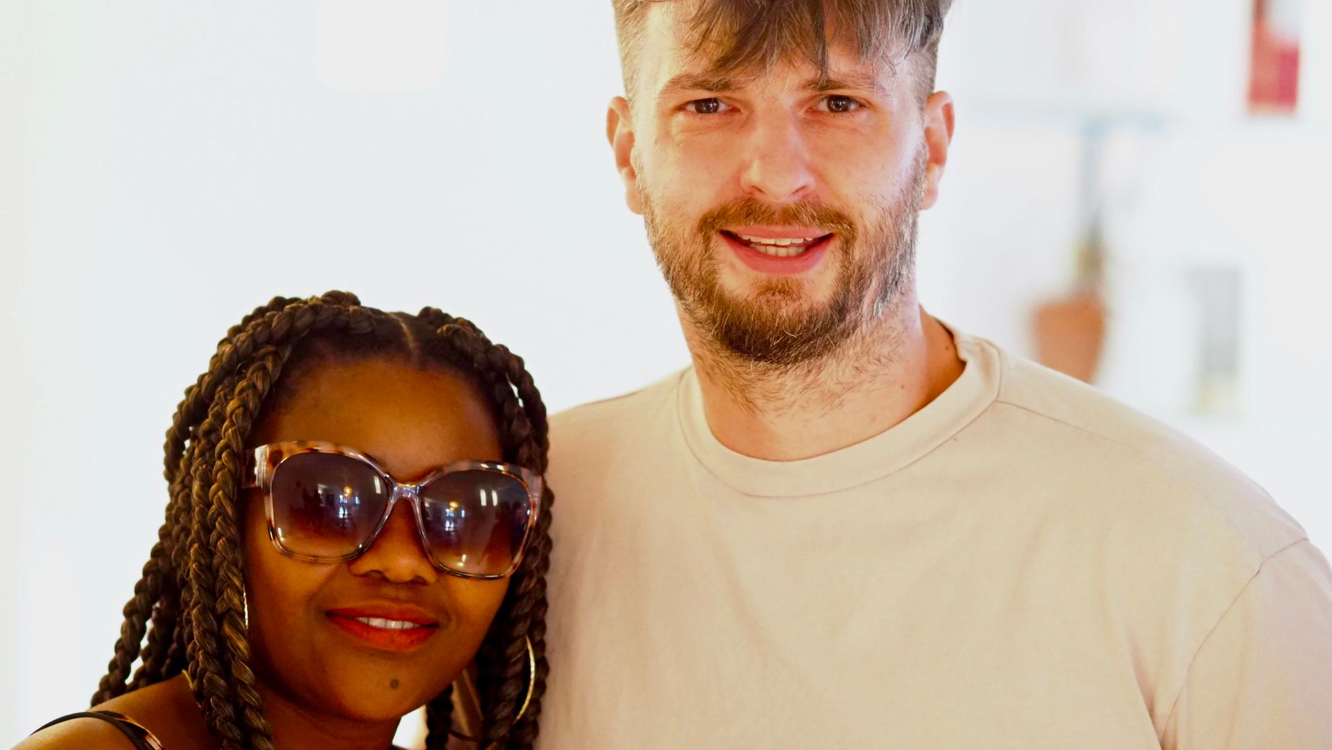 Two smiling people posing together indoors, one wearing sunglasses, in warm bright light