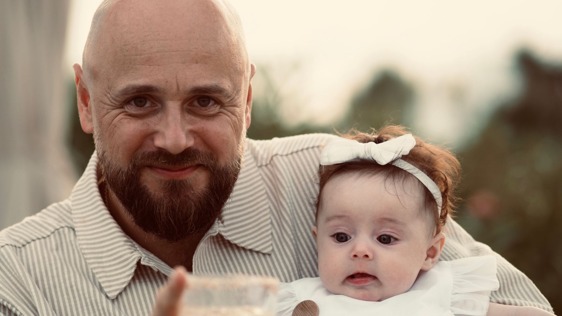 Man holding a baby with a white headband, both facing the camera outdoors with a soft blurred background