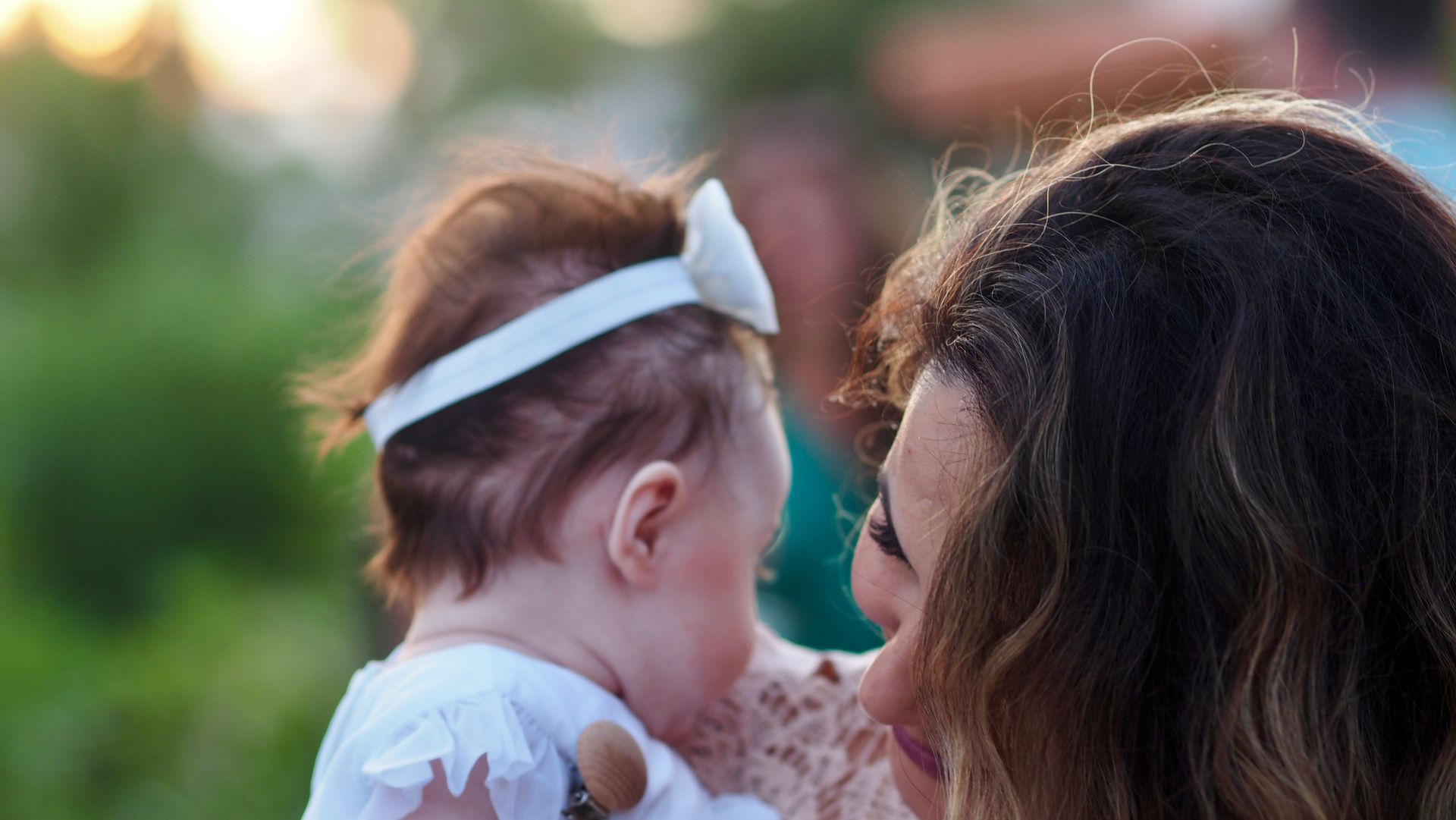 Woman facing a baby with a white bow headband outdoors, both in soft evening light