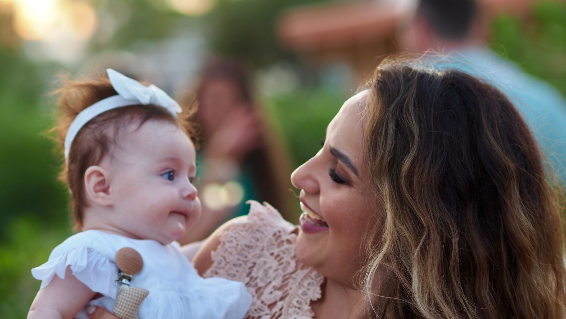 Woman smiling at a baby in a white outfit and bow, outdoors with blurred greenery in the background
