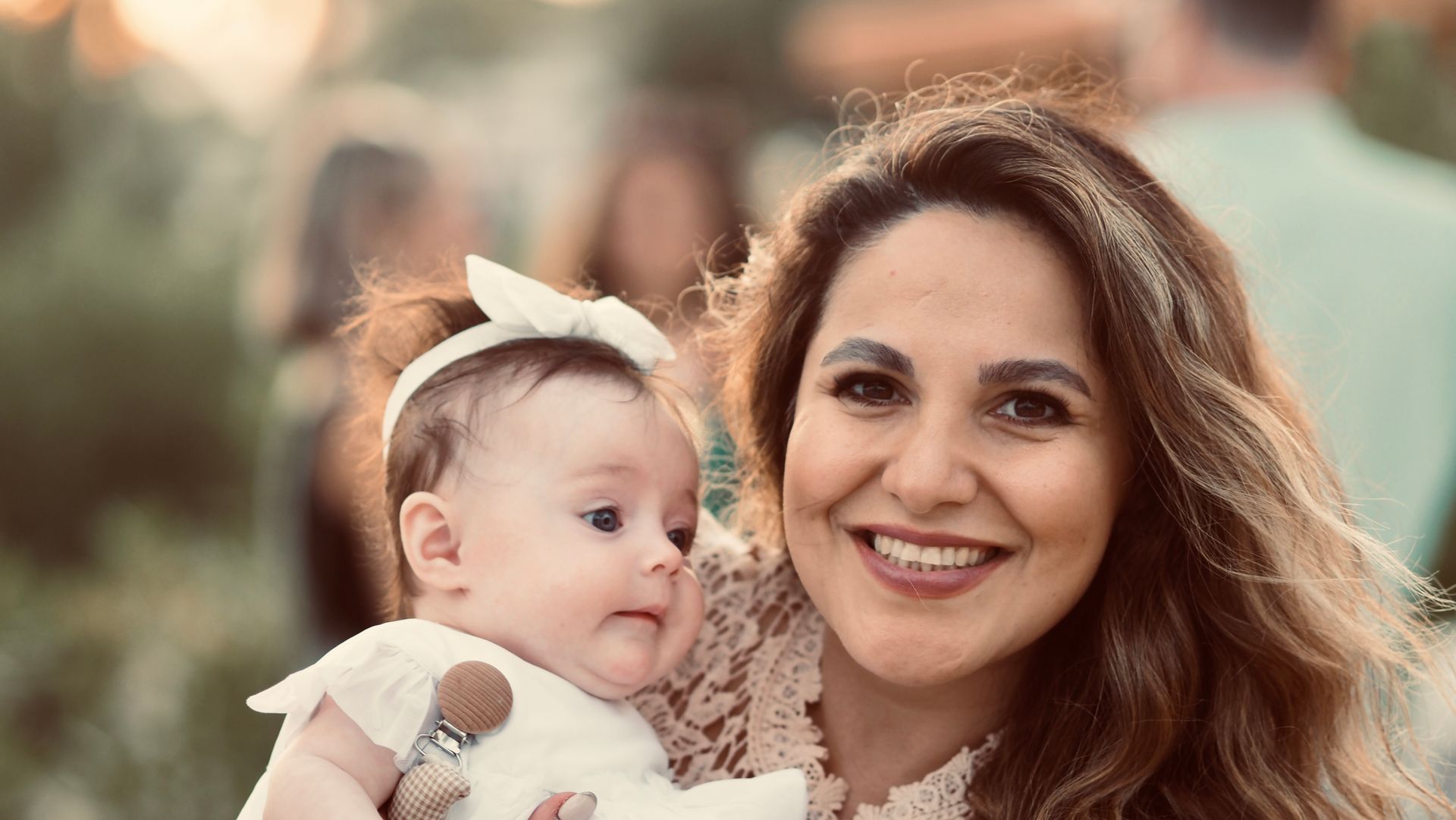 Smiling woman holding a baby outdoors, with warm sunlight and a soft blurred background