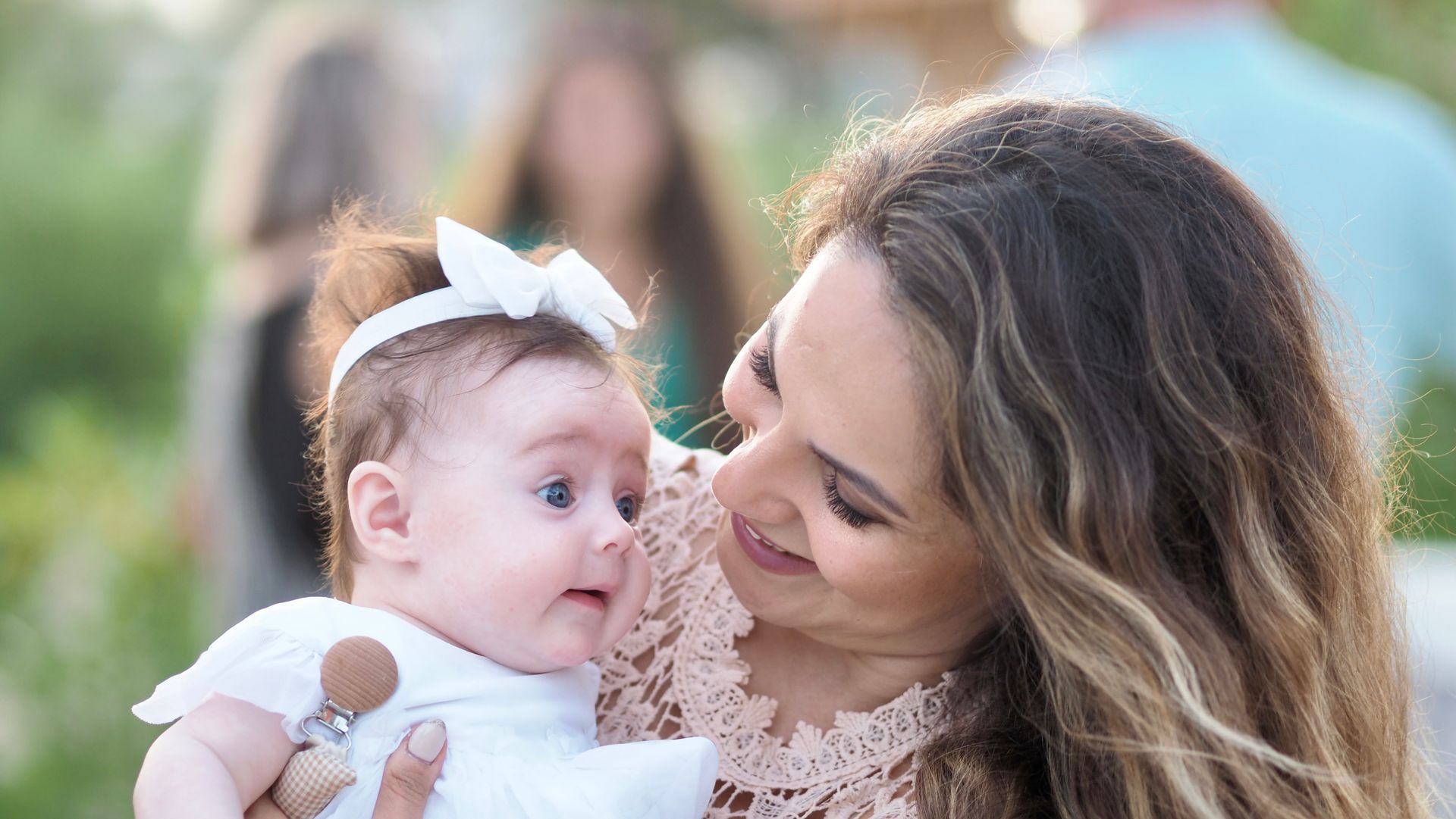 Woman holding a baby with a white bow, smiling outdoors with a blurred green background