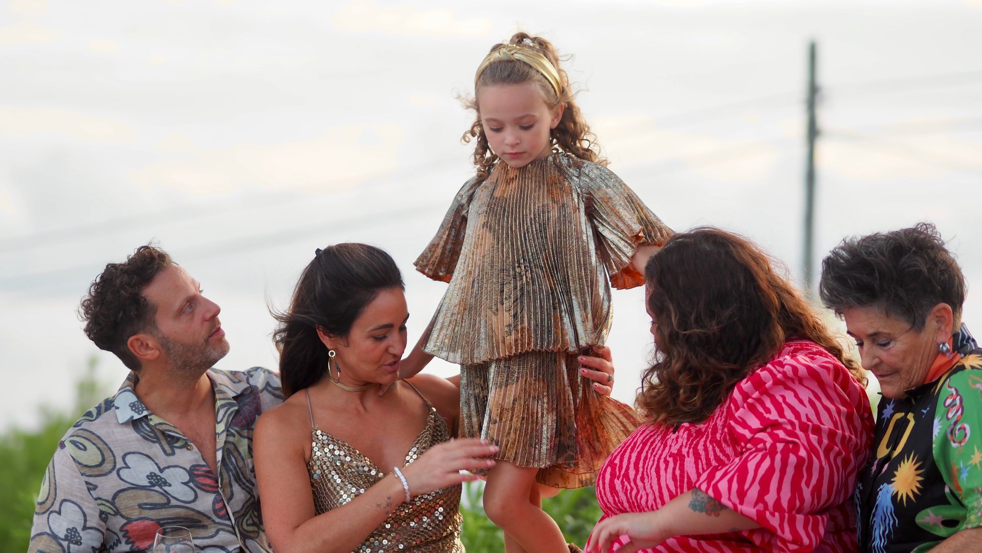 Small child in straw costume standing on adults’ laps outdoors, with smiling family around her