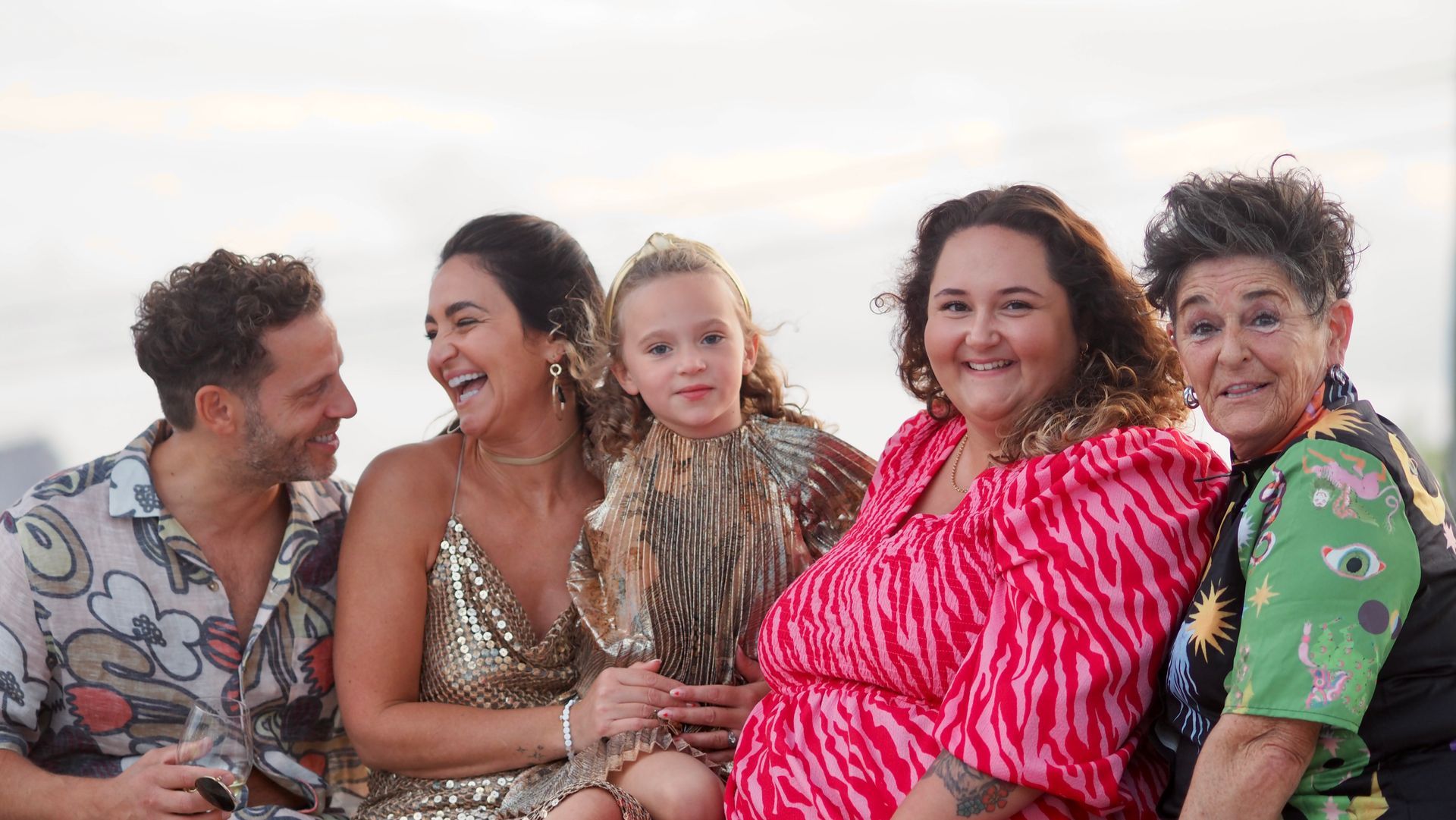 Five people sitting closely together on a couch, smiling and laughing in colorful patterned clothes.